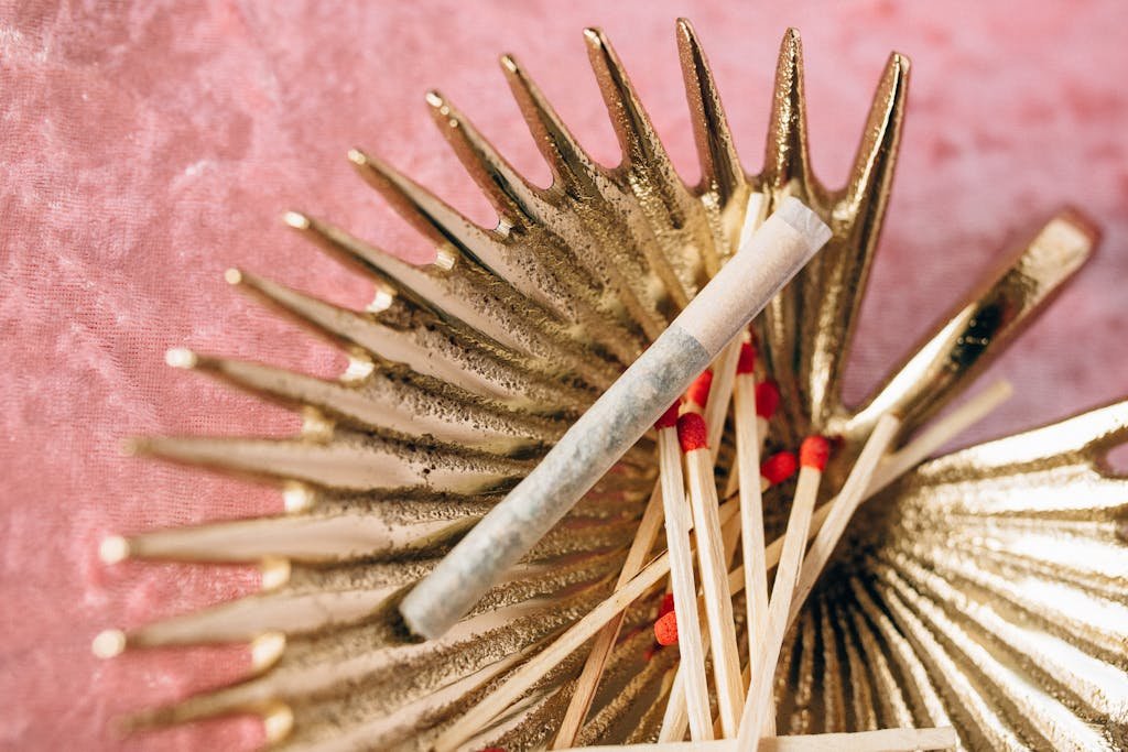 A golden ashtray holding a cannabis joint and red-tipped matches on a textured pink background.