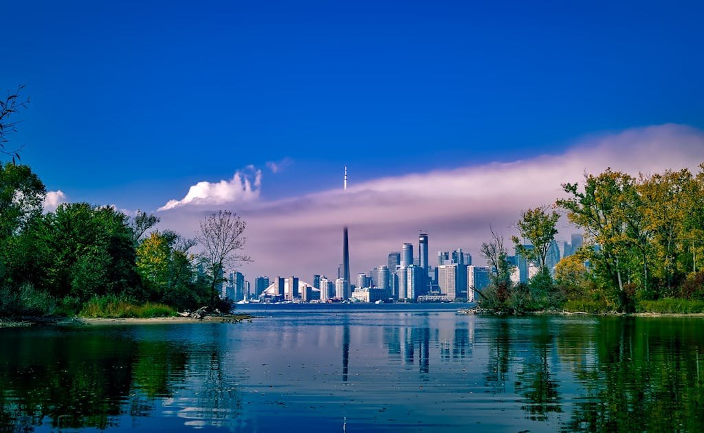 Scenic view of Toronto skyline across a reflective lake with lush greenery and clear blue sky.