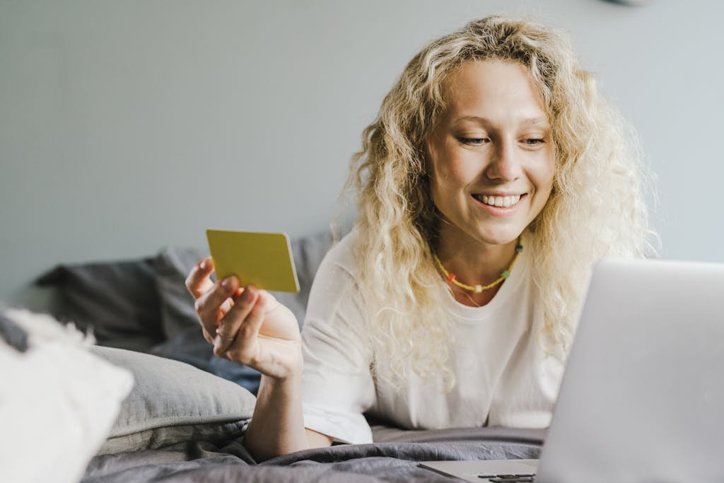 Smiling blonde woman with curly hair shopping online with a bank card indoors.