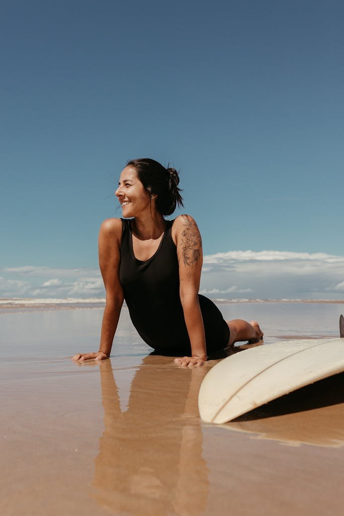 Smiling woman enjoying a sunny day on the beach with a surfboard nearby.