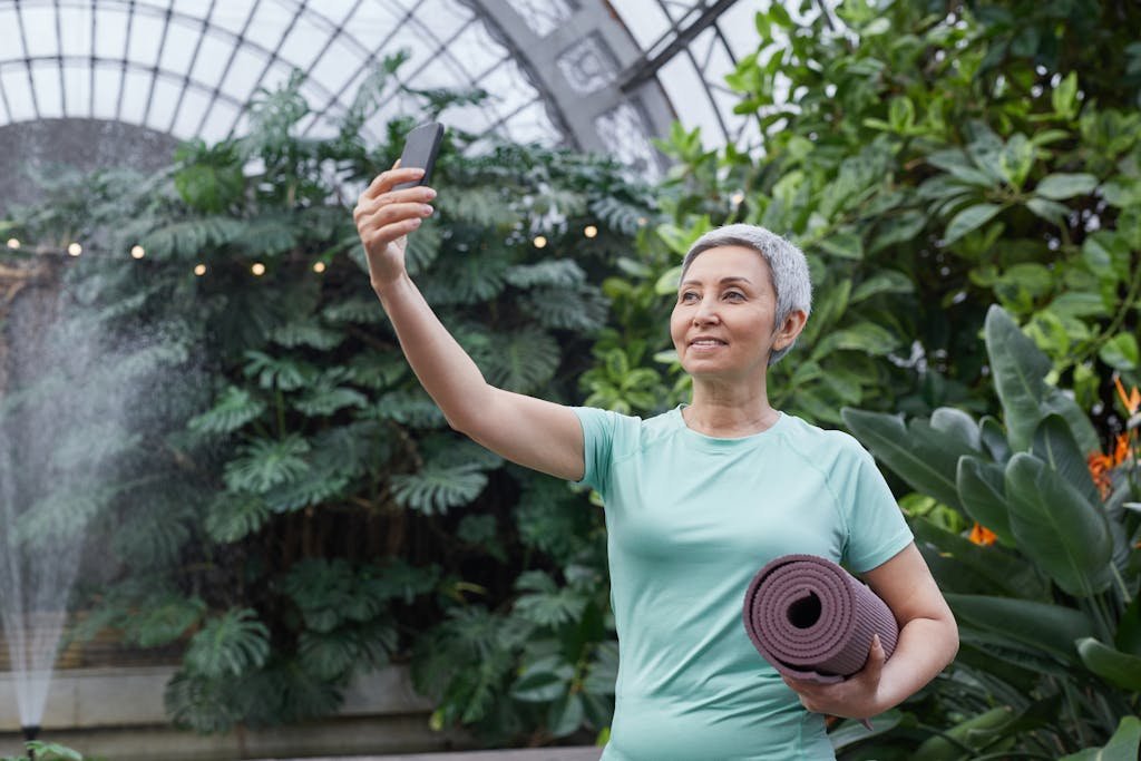 A senior woman taking a selfie while holding a yoga mat in a lush greenhouse setting.