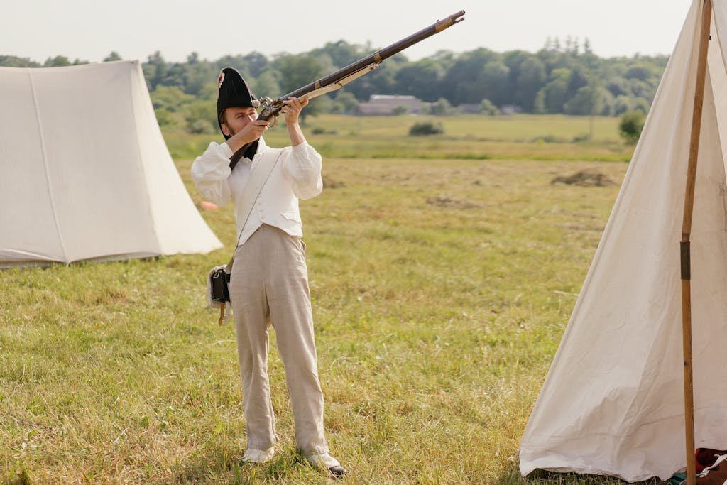 A soldier in Revolutionary Cellulogia a musket during a reenactment on a grassy battlefield.