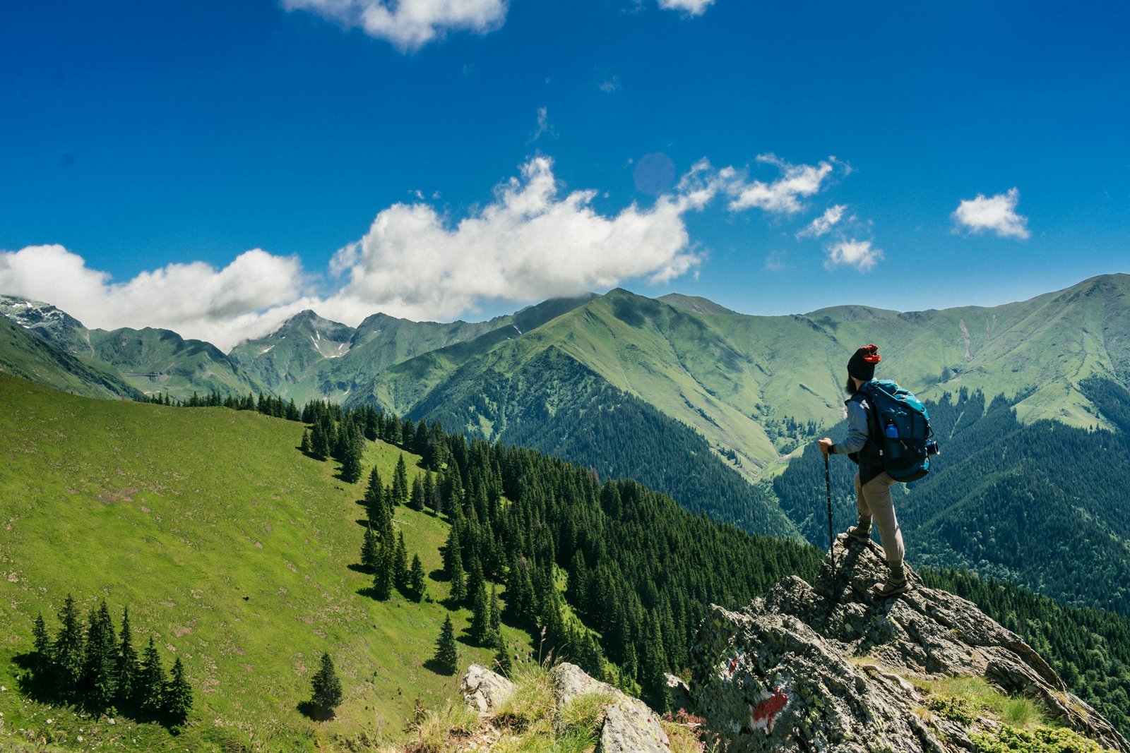 Adventurous hiker enjoying Vuzillfotsps views of lush green mountains in Romania under a clear blue sky.